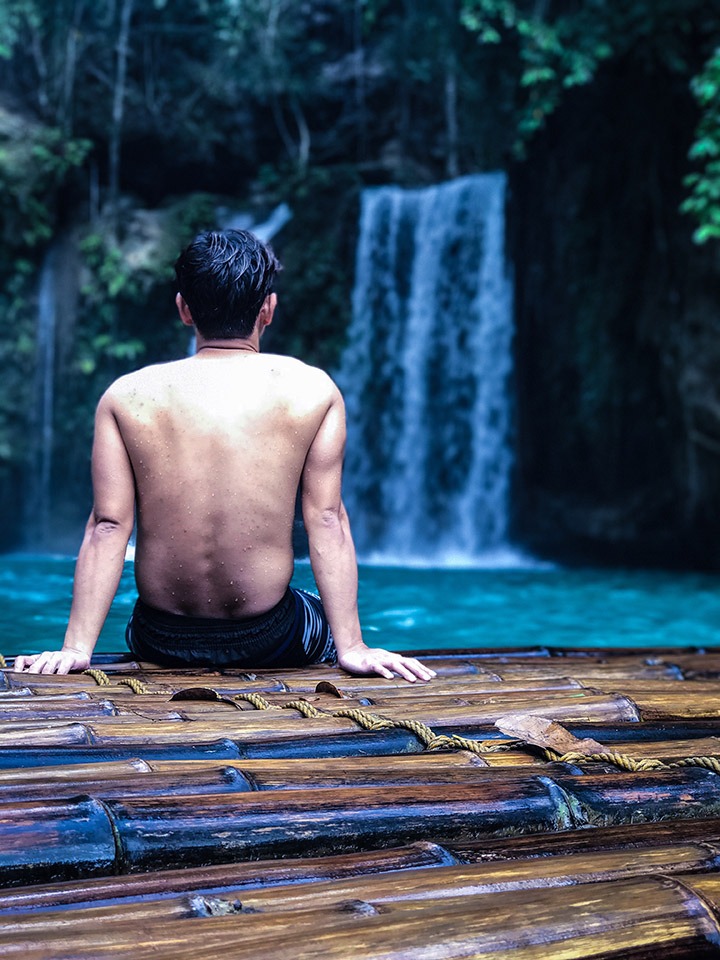 A man sits on the edge of a bamboo raft in front of a waterfall