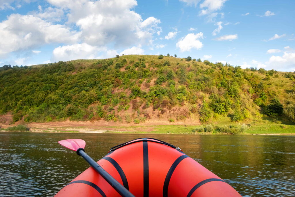 A point-of-view shot from within an orange raft. The camera looks out at still water and rolling hills.