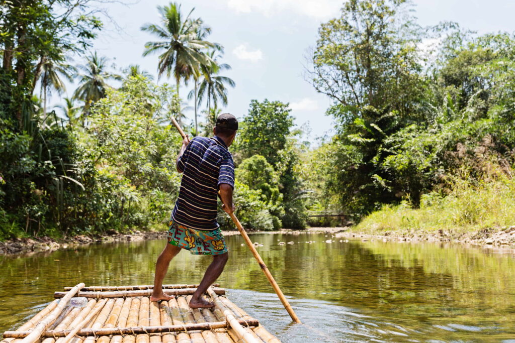 A man steers a bamboo raft on a river using a bamboo pole. 